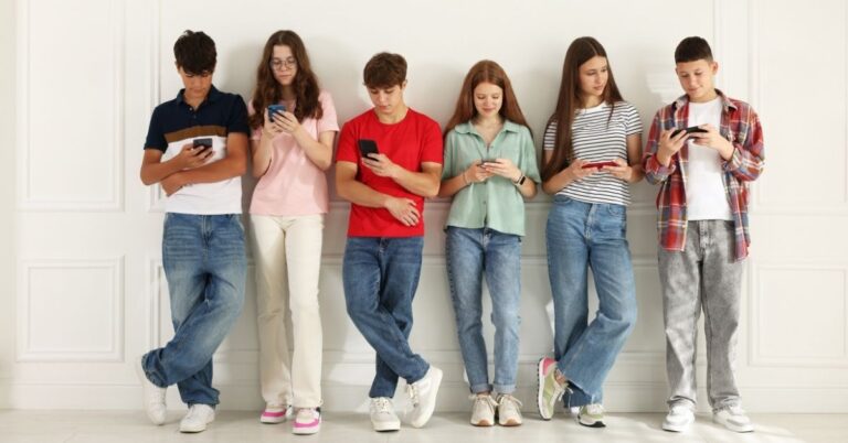 Six teenagers standing against a wall while looking at their phones and tablets.