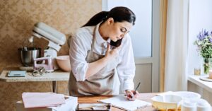 Small business owner writing notes while talking on the phone in a bright workspace with baking supplies nearby.