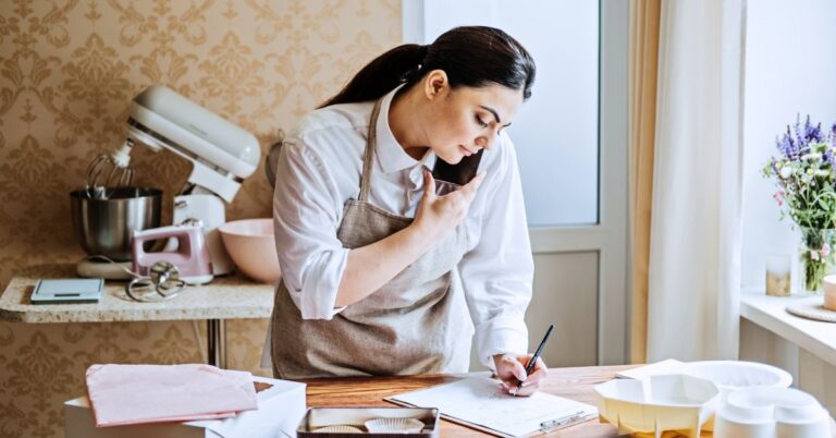 Small business owner writing notes while talking on the phone in a bright workspace with baking supplies nearby.