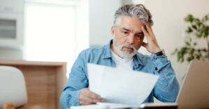 Concerned man sitting at a table and reviewing documents beside a laptop.