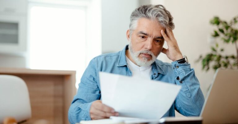 Concerned man sitting at a table and reviewing documents beside a laptop.