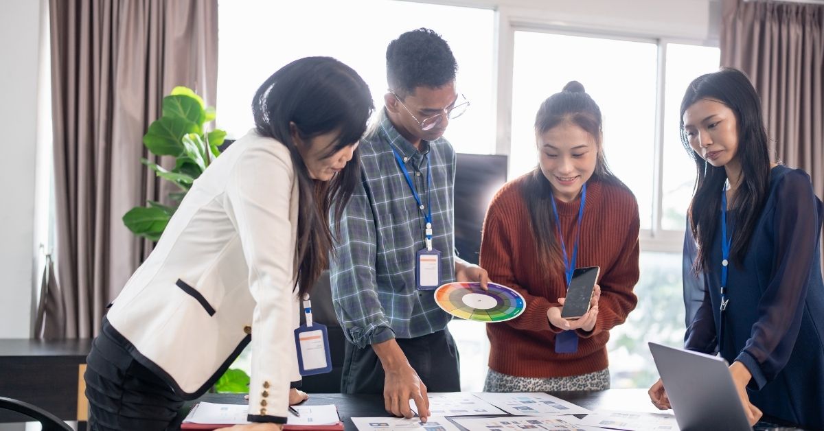 A UX design team reviewing mobile app mockups, a color palette, and a smartphone during a collaborative design session.