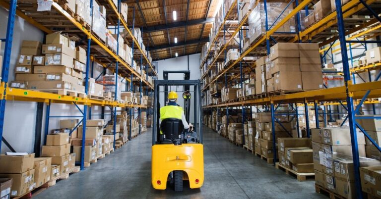 Forklift operator driving through a warehouse aisle lined with tall shelves and stacked boxes.