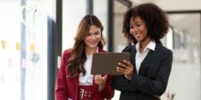 Is digital marketing a good career? Salary, demand, and fit Two business professionals smiling while looking at a tablet together in a bright office hallway.