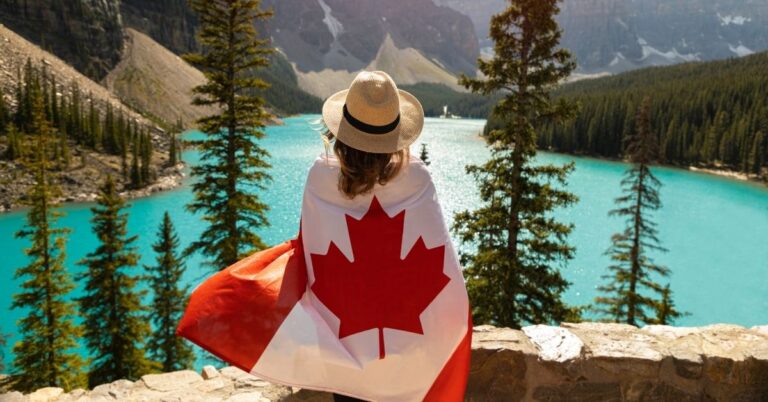 Person wrapped in a Canadian flag looking out over a bright turquoise mountain lake surrounded by trees.