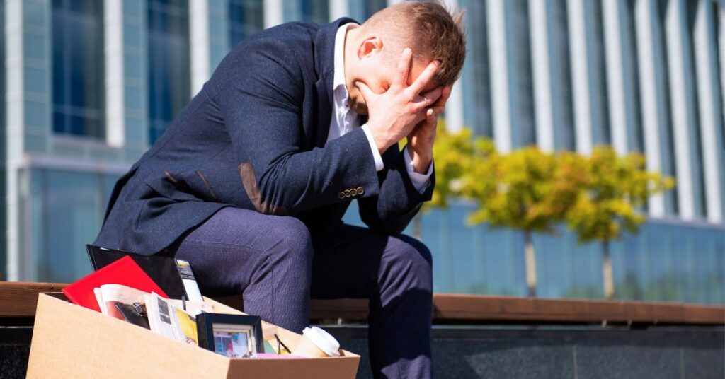 Distressed business professional sitting beside a box of office belongings outside a corporate building.