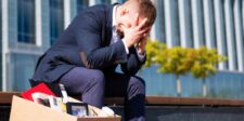 Distressed business professional sitting beside a box of office belongings outside a corporate building.