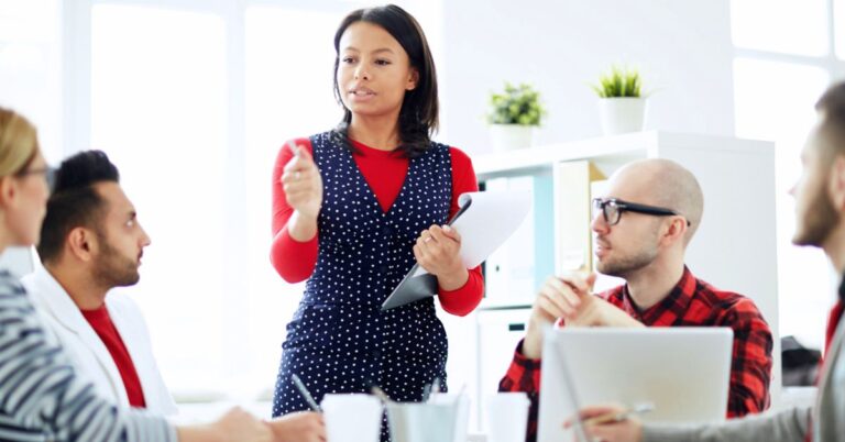Woman leading a business meeting while holding papers as colleagues sit around a table with laptops.