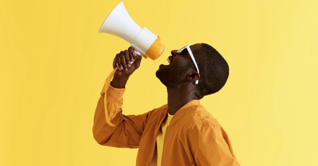 Man speaking into a megaphone against a bright yellow background.