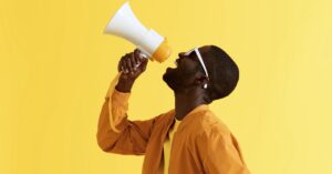 Man speaking into a megaphone against a bright yellow background.