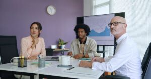 Three coworkers in a meeting looking toward a presentation screen showing business performance charts.