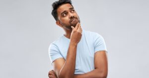 Man in a light blue shirt looking up with one hand on his chin against a gray background.