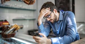 Concerned man in glasses looking at his smartphone while leaning on a counter.