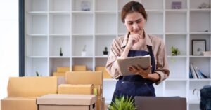 Small business owner reviewing a clipboard beside shipping boxes and a laptop in a workspace.