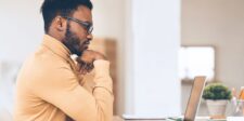 A man in glasses thinking while looking at a laptop.