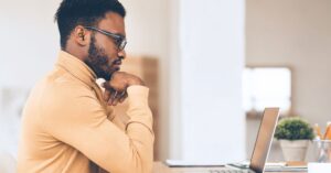A man in glasses thinking while looking at a laptop.