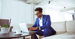 Man in a blue suit working on a laptop while seated on a couch in a bright modern room.