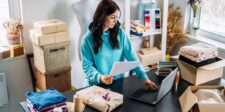 Woman working at a laptop while reviewing papers at a table surrounded by folded clothing, packages, and shipping supplies.