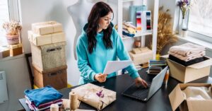 Woman working at a laptop while reviewing papers at a table surrounded by folded clothing, packages, and shipping supplies.