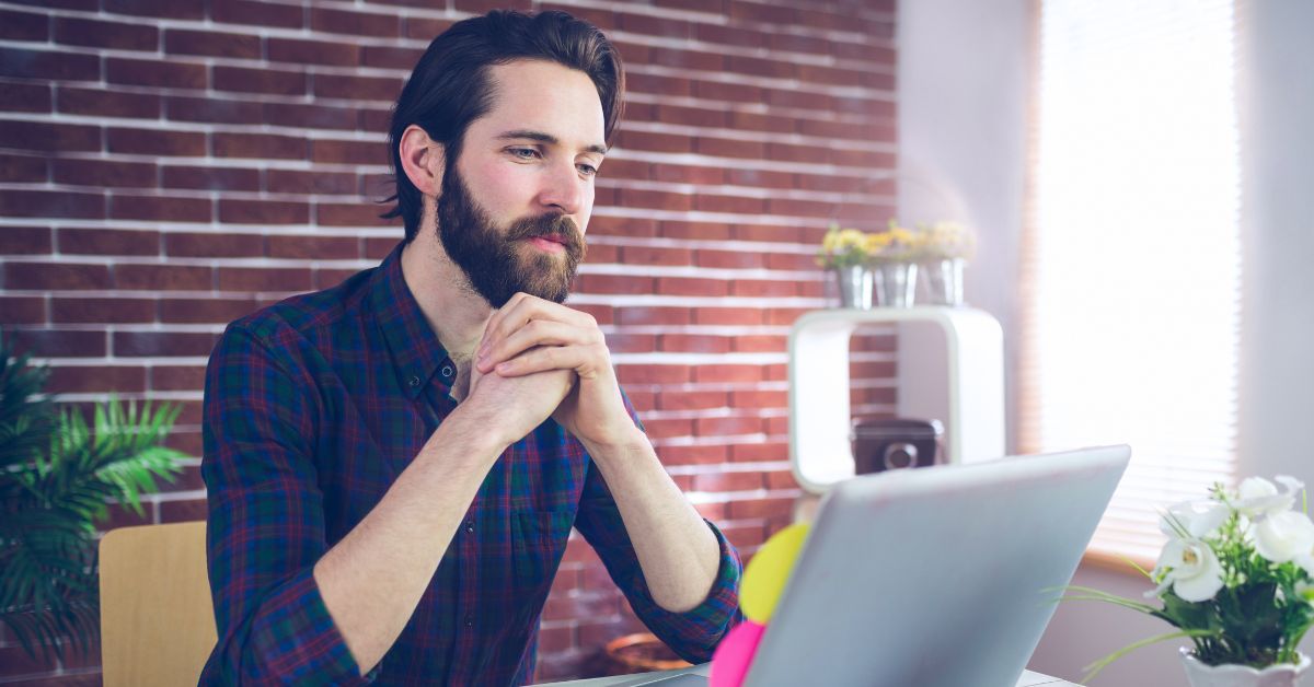 Bearded man sitting at a desk and looking at a laptop in a bright office.