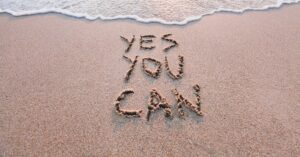 Words reading “Yes You Can” written in sand near the edge of the water.
