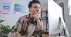 Man with headphones around his neck looking thoughtfully at a desktop computer in a creative workspace.