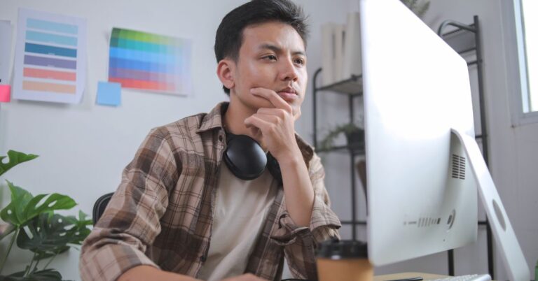 Man with headphones around his neck looking thoughtfully at a desktop computer in a creative workspace.
