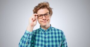 Man wearing glasses and a green plaid shirt looking toward the camera with a skeptical expression.