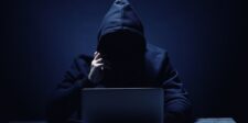 Hooded figure sitting behind a laptop in a dark setting, representing a cyberattack or security threat.