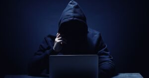 Hooded figure sitting behind a laptop in a dark setting, representing a cyberattack or security threat.
