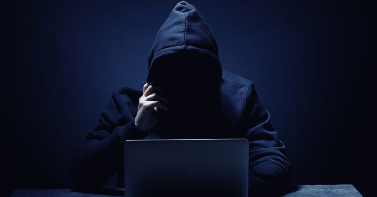 Hooded figure sitting behind a laptop in a dark setting, representing a cyberattack or security threat.
