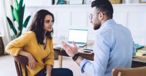 Two business professionals discussing ideas across a table with a laptop in the background.