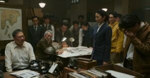 Film characters gathered around a table in a dimly lit office scene from Godzilla Minus Zero.