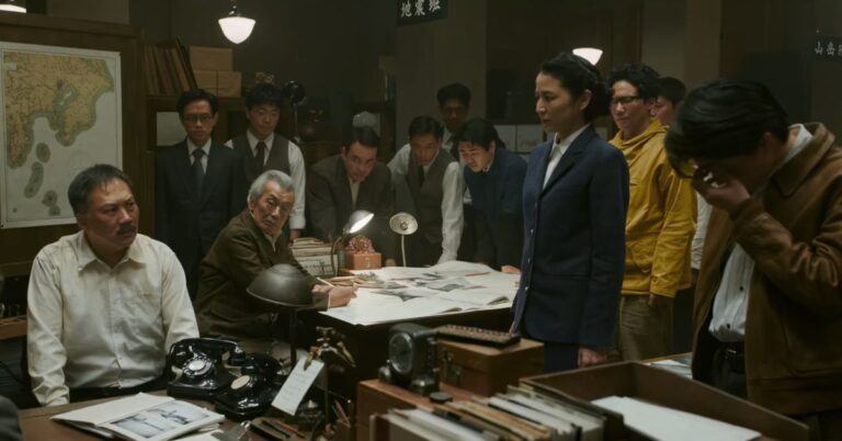 Film characters gathered around a table in a dimly lit office scene from Godzilla Minus Zero.