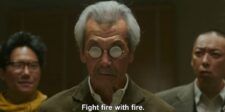Older man wearing round reflective glasses standing between two men in a dramatic film scene.