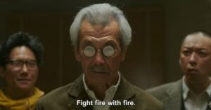 Older man wearing round reflective glasses standing between two men in a dramatic film scene.