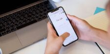 Google Ads AI Max auto-upgrades are rolling out now Person using Google Search on a smartphone beside an open laptop.