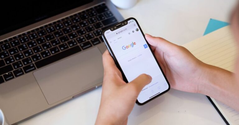 Person using Google Search on a smartphone beside an open laptop.