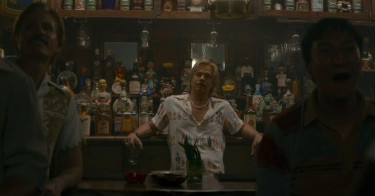 Man seated at a dimly lit bar surrounded by bottles and vintage decorations in a film scene.