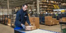 Holiday shipping costs are moving from forecast to invoice Warehouse worker handling parcels on a conveyor in a busy fulfilment centre.