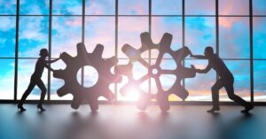 Silhouettes of two businesspeople pushing interlocking gears in front of large windows at sunset, representing technical systems working together.
