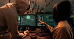 Two hooded people reviewing code and system activity across multiple computer monitors.