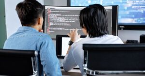 Two cybersecurity professionals reviewing code and system activity on multiple computer screens.
