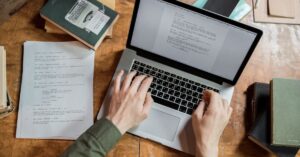Overhead view of a person typing on a MacBook Pro at a wooden table with books and papers.
