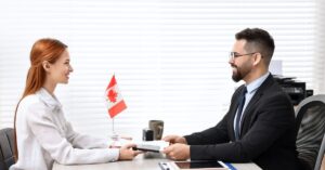 Job candidate and interviewer passing a document across a desk with a small Canadian flag