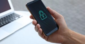 A hand holds a smartphone displaying an open padlock symbol beside a laptop.