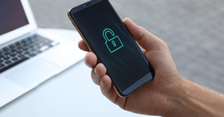 A hand holds a smartphone displaying an open padlock symbol beside a laptop.