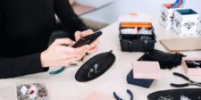 Etsy tightens AI listing rules but keeps seller-prompted AI art Person holding a smartphone above a worktable with handmade jewelry, tools, and boxes.