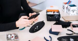 Person holding a smartphone above a worktable with handmade jewelry, tools, and boxes.