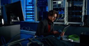 IT administrator working on a laptop beside server racks and network equipment.